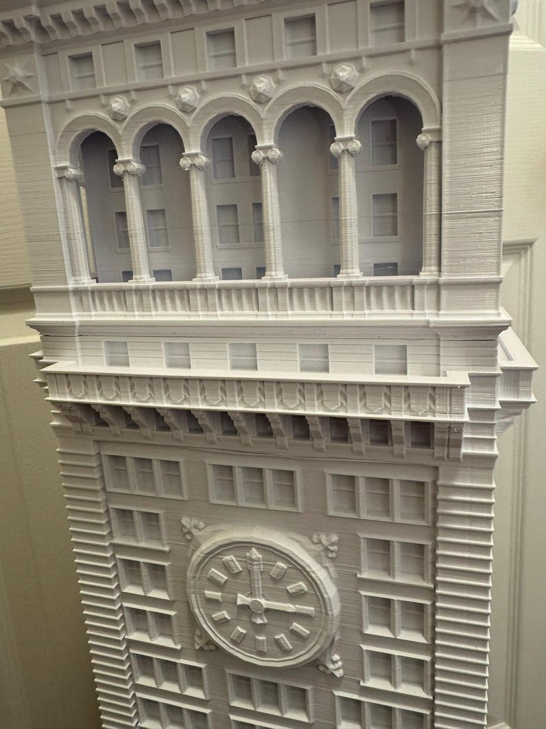 Op de afbeelding: Een wit 3D-geprint model van een hoog gebouw met een klok op de voorgevel. Het gebouw heeft veel ramen en een decoratieve kroonlijst.
