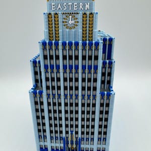 May include: A light blue model of a building with the word "EASTERN" on top. The building has a clock and gold and blue accents. The windows are black and the building is on a white surface.