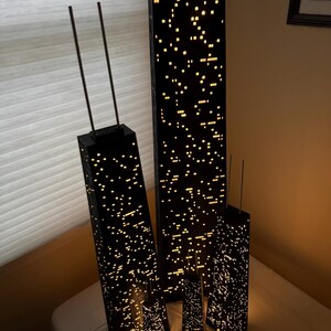May include: A collection of black, illuminated skyscraper models with a city lights design. The models vary in size, with two taller structures and several smaller ones. The lights create a warm, inviting glow, suggesting a nighttime cityscape.