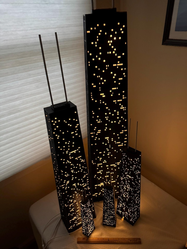 May include: A collection of black, illuminated skyscraper models with a city lights design. The models vary in size, with two taller structures and several smaller ones. The lights create a warm, inviting glow, suggesting a nighttime cityscape.