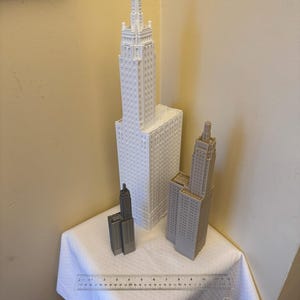 May include: Three white and grey miniature building models on a white tablecloth. The tallest building is white and has a pointed roof. The other two buildings are grey and have flat roofs. A ruler is visible in the foreground.