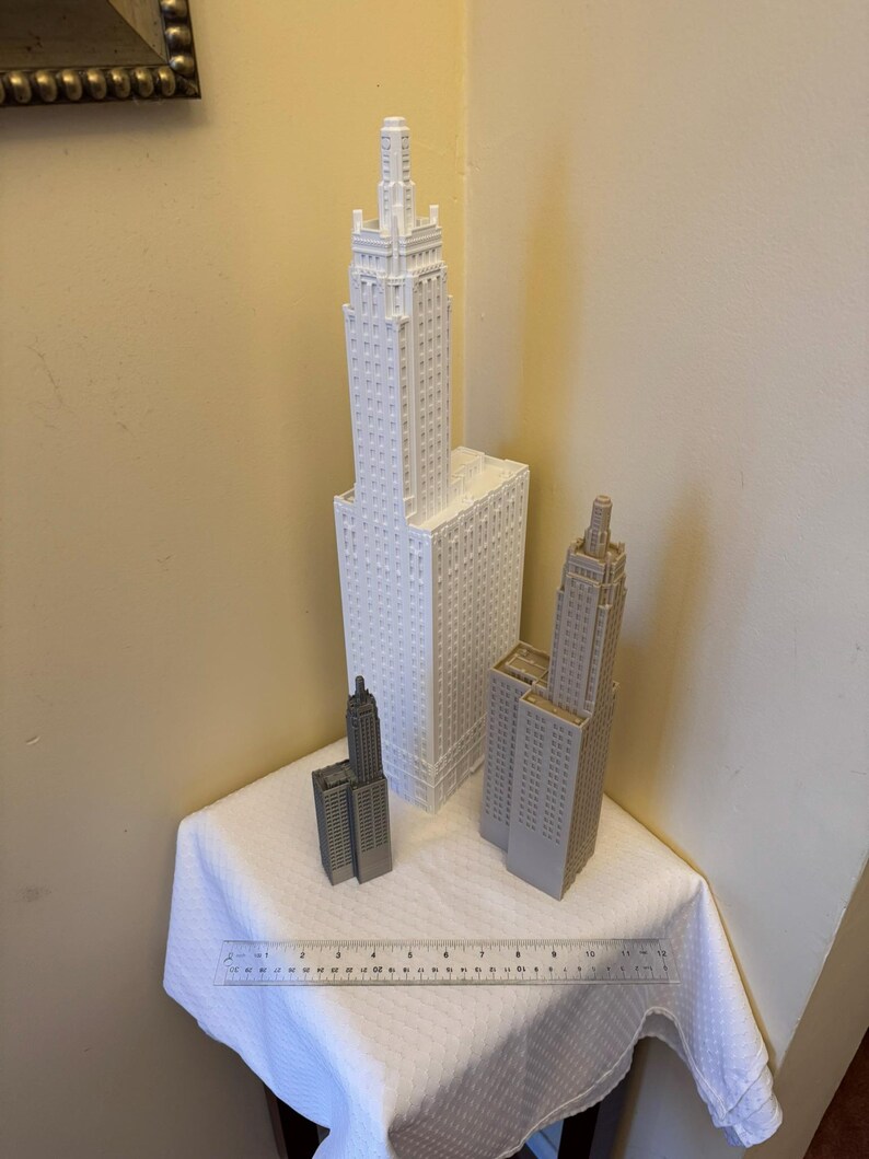 May include: Three white and grey miniature building models on a white tablecloth. The tallest building is white and has a pointed roof. The other two buildings are grey and have flat roofs. A ruler is visible in the foreground.
