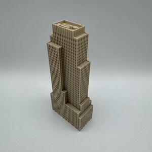 May include: A beige-colored miniature skyscraper model with a multi-tiered design. The building has numerous small, square windows and a flat roof. The model is a detailed representation of a tall building.