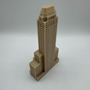 Może przedstawiać: Beżowy miniaturowy model wieżowca ze szczeg&oacute;łową fasadą, z wieloma prostokątnymi oknami i warstwowym projektem. Model ma wysoką centralną wieżę i kilka niższych sekcji.