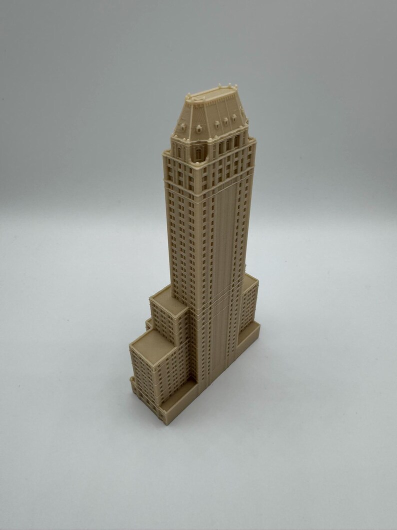 Może przedstawiać: Beżowy miniaturowy model wieżowca ze szczeg&oacute;łową fasadą, z wieloma prostokątnymi oknami i warstwowym projektem. Model ma wysoką centralną wieżę i kilka niższych sekcji.