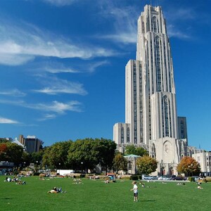 May include: A large, white stone building with a tall spire, surrounded by green grass and trees. People are walking and sitting on the grass.
