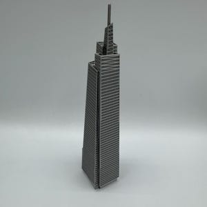 May include: A gray metal model of a skyscraper with a tall antenna. The building has many windows and a detailed architectural design. The model is standing on a white surface.