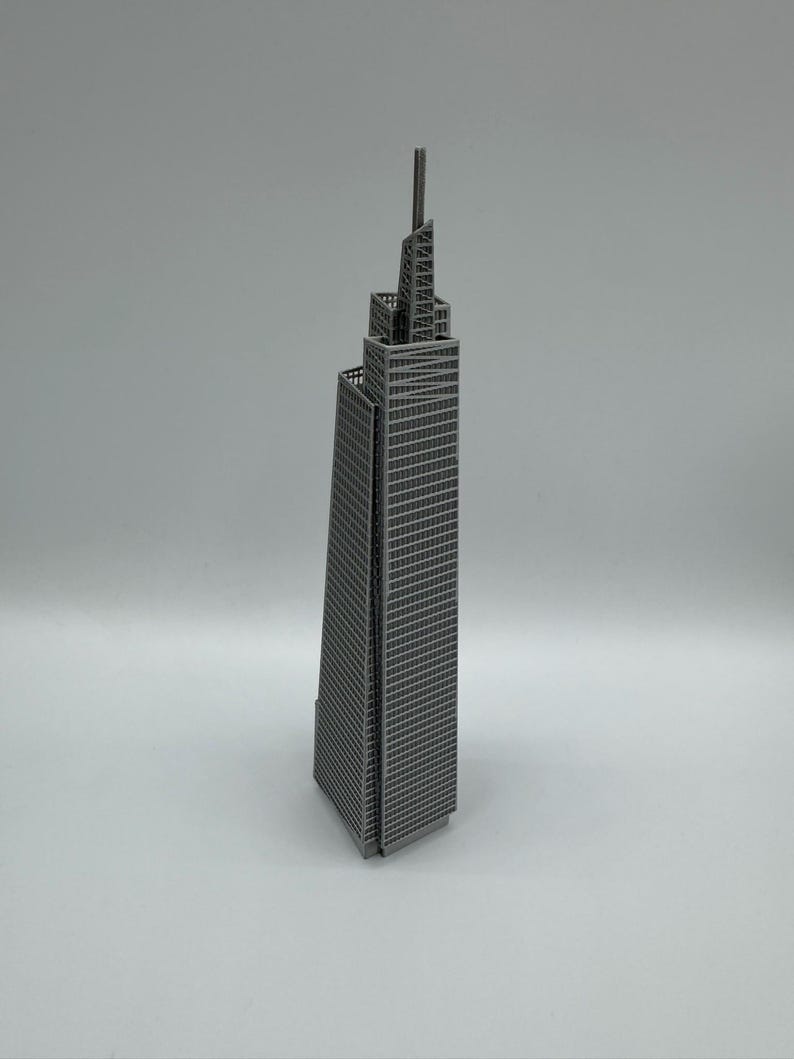May include: A gray metal model of a skyscraper with a tall antenna. The building has many windows and a detailed architectural design. The model is standing on a white surface.