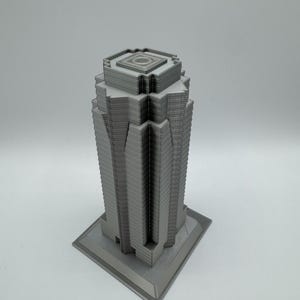 May include: A gray, 3D-printed model of a skyscraper. The building has a rectangular base and a multi-tiered design, with a detailed facade. The top of the building features a square, recessed design. The model is set against a plain white background.