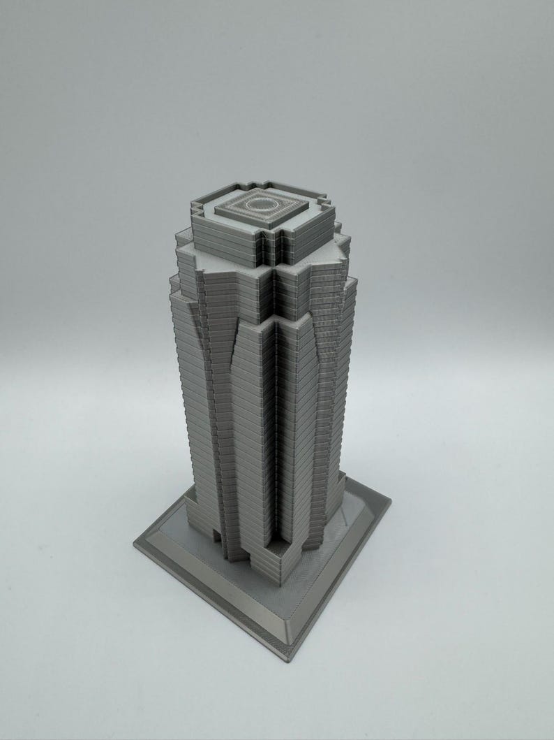 May include: A gray, 3D-printed model of a skyscraper. The building has a rectangular base and a multi-tiered design, with a detailed facade. The top of the building features a square, recessed design. The model is set against a plain white background.