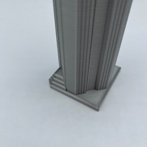 May include: A gray 3D printed model of a tall building with a square base and a set of stairs leading up to the main structure.