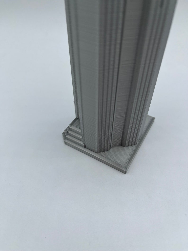 May include: A gray 3D printed model of a tall building with a square base and a set of stairs leading up to the main structure.