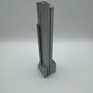 May include: A gray, 3D-printed model of a skyscraper. The building has a rectangular base with a tall, slender tower. The tower features numerous windows and a flat roof. The model is a solid gray color and sits on a white surface.