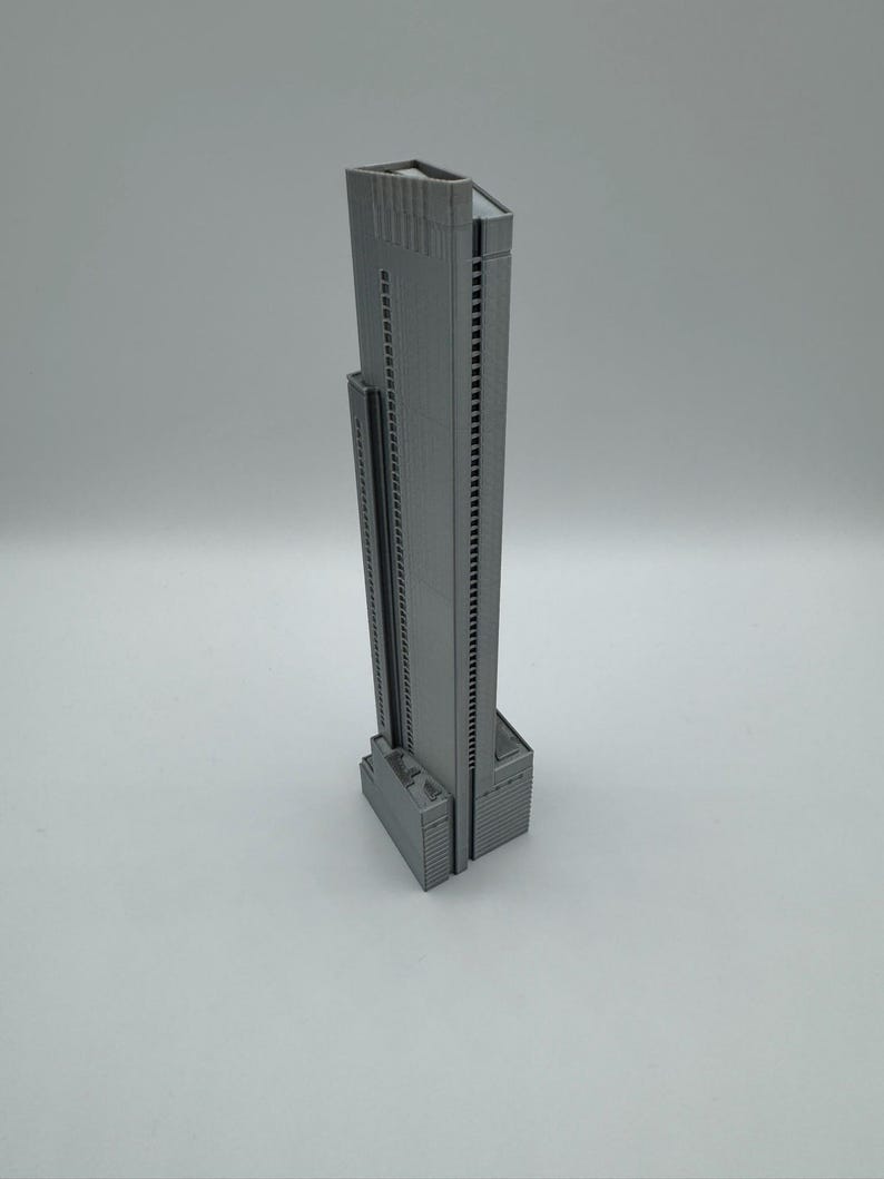 May include: A gray, 3D-printed model of a skyscraper. The building has a rectangular base with a tall, slender tower. The tower features numerous windows and a flat roof. The model is a solid gray color and sits on a white surface.