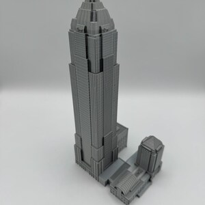 May include: A gray, 3D-printed model of a skyscraper with a detailed design. The building has a tall, tiered structure with a pointed top and a smaller adjacent building. The model is on a white surface.