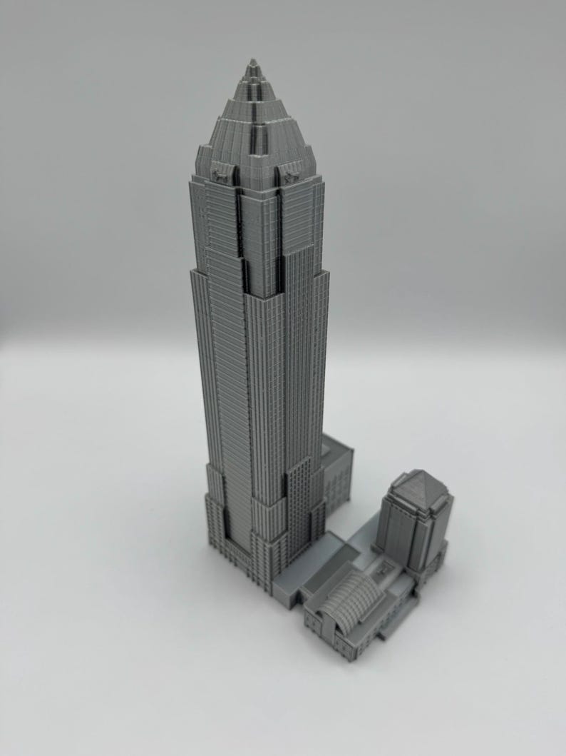 May include: A gray, 3D-printed model of a skyscraper with a detailed design. The building has a tall, tiered structure with a pointed top and a smaller adjacent building. The model is on a white surface.