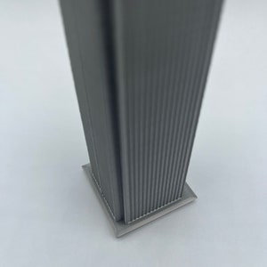 Op de afbeelding: Een grijs, 3D-geprint model van een wolkenkrabber met een vierkante basis. Het model heeft een getextureerd oppervlak en is ontworpen om eruit te zien als een modern gebouw.