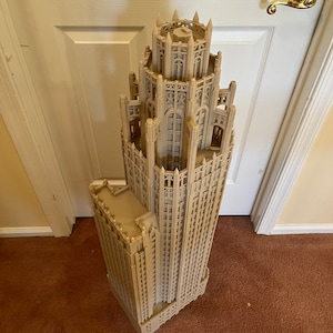 May include: A detailed 3D printed model of a tall, ornate building with a pointed roof and many windows. The model is made of a light brown plastic and is standing on a brown carpet.