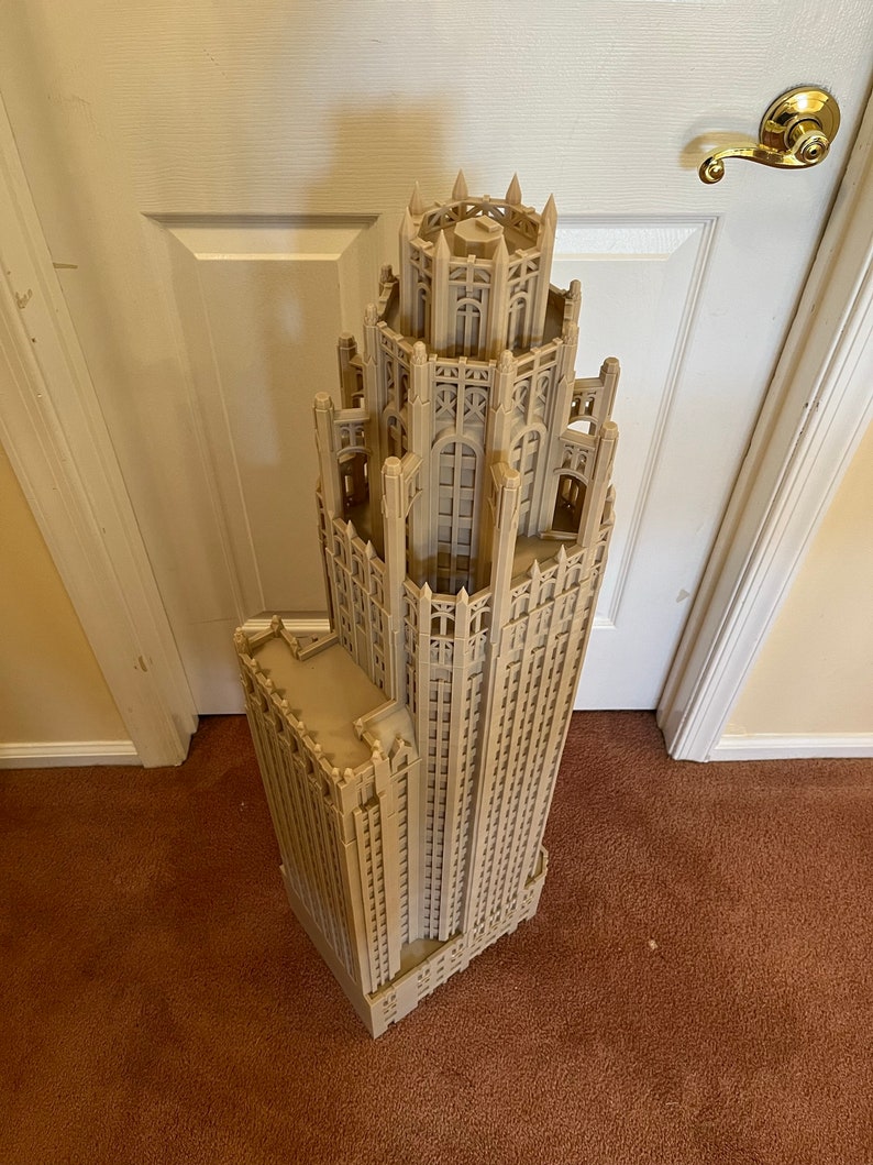 May include: A detailed 3D printed model of a tall, ornate building with a pointed roof and many windows. The model is made of a light brown plastic and is standing on a brown carpet.