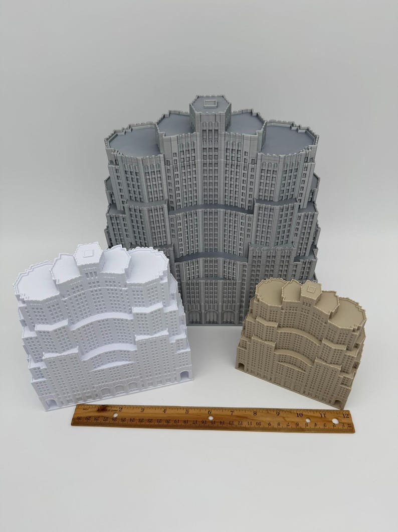 May include: Three miniature building models in grey, white, and tan, with a wooden ruler. The buildings have detailed facades with windows and architectural features. The ruler is approximately 30 cm long.