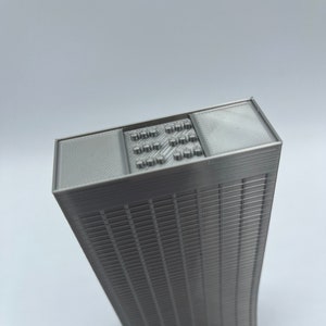 May include: A 3D printed model of a skyscraper with a detailed facade and a flat roof with a grid pattern.
