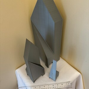 May include: Three gray geometric shapes made of 3D printed plastic. The shapes are arranged on a white tablecloth with a ruler in the foreground.