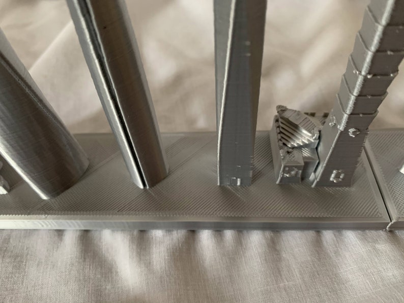 May include: A collection of gray 3D-printed architectural models, including cylindrical, twisted, and arched structures, are displayed on a textured base. The models showcase various geometric shapes and designs, highlighting the potential of 3D printing for architectural applications.