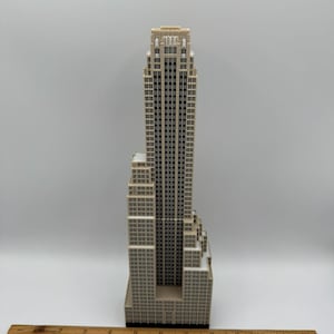 May include: A beige and black miniature skyscraper model with a detailed facade of windows. The model is approximately 10 inches tall and has a ruler placed at the base for scale.