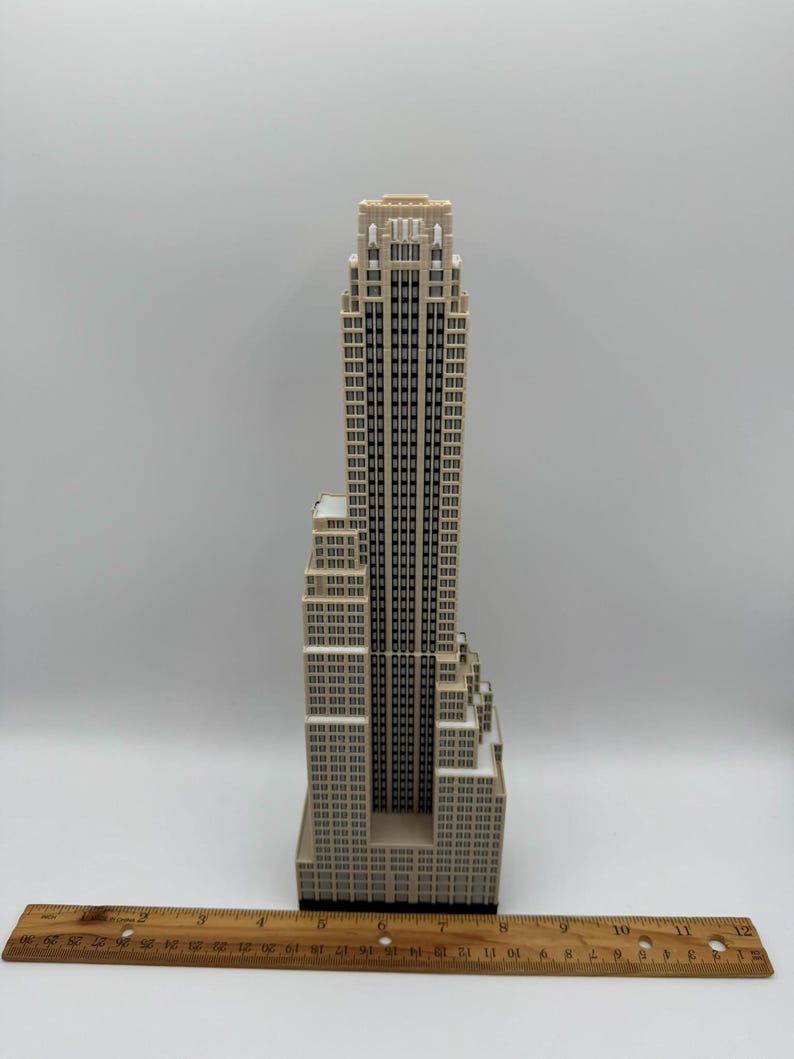 May include: A beige and black miniature skyscraper model with a detailed facade of windows. The model is approximately 10 inches tall and has a ruler placed at the base for scale.