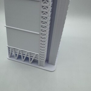 May include: A 3D-printed model of a modern skyscraper. The building is primarily white with detailed architectural features, including a series of repeating geometric shapes along the side and a support structure at the base. The model is set against a plain white background.