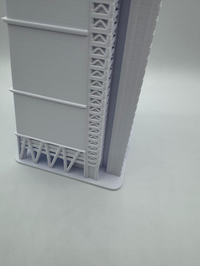May include: A 3D-printed model of a modern skyscraper. The building is primarily white with detailed architectural features, including a series of repeating geometric shapes along the side and a support structure at the base. The model is set against a plain white background.