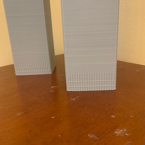 May include: Two grey 3D printed models of skyscrapers, one slightly taller than the other, on a wooden surface.