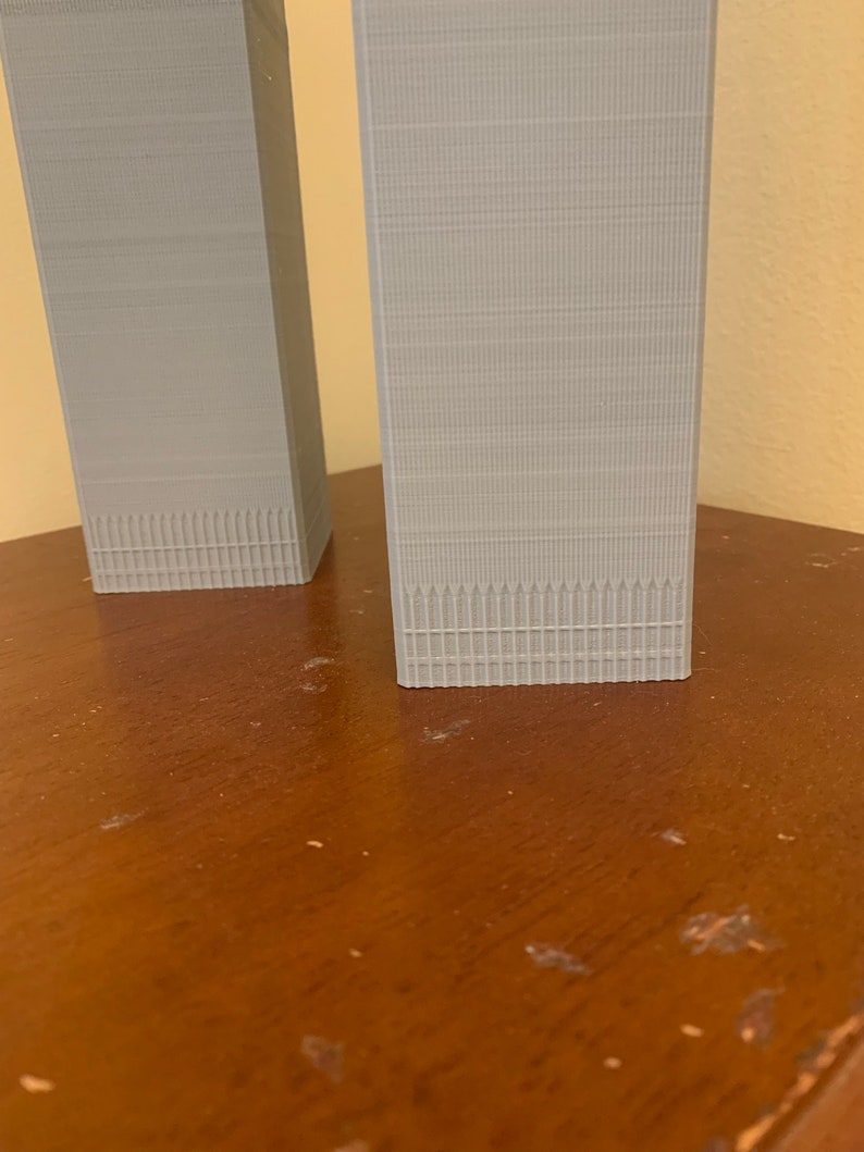 May include: Two grey 3D printed models of skyscrapers, one slightly taller than the other, on a wooden surface.