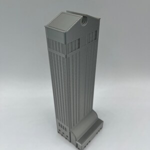May include: A gray 3D printed model of a tall building with a flat roof and a small window on the top. The building has a series of windows along the sides and a small entrance at the bottom.