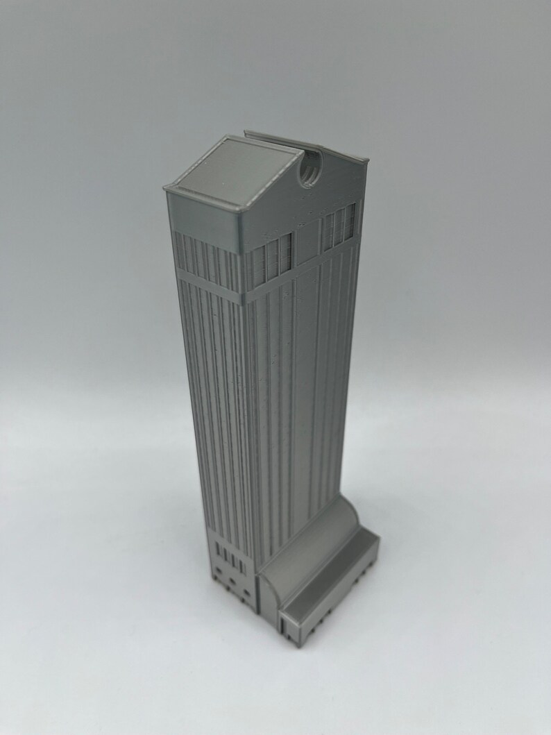 May include: A gray 3D printed model of a tall building with a flat roof and a small window on the top. The building has a series of windows along the sides and a small entrance at the bottom.