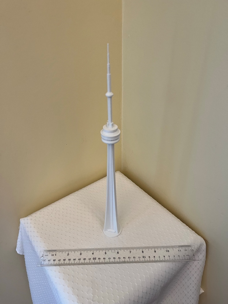 May include: A white 3D-printed model of the CN Tower, a tall structure with a spire, standing on a white surface. A clear ruler with centimeter markings is placed in front of the model. The tower is a prominent landmark.