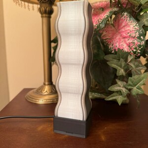 K&ouml;nnte beinhalten: Eine dekorative Lampe mit einem wellenf&ouml;rmigen, vertikalen Design. Die Lampe hat eine hellgraue Mitte mit dunkelgrauen Kanten und einen schwarzen Sockel. Die Lampe ist an eine Steckdose angeschlossen. Die Lampe steht auf einer Holzoberfl&auml;che.