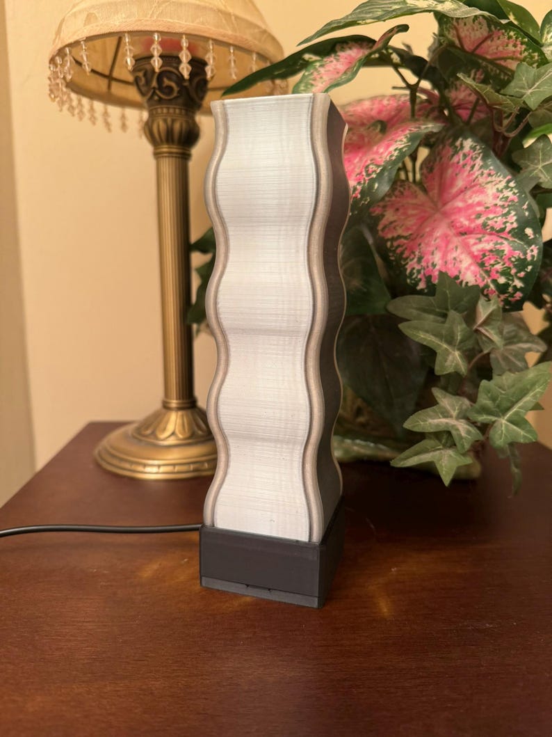 K&ouml;nnte beinhalten: Eine dekorative Lampe mit einem wellenf&ouml;rmigen, vertikalen Design. Die Lampe hat eine hellgraue Mitte mit dunkelgrauen Kanten und einen schwarzen Sockel. Die Lampe ist an eine Steckdose angeschlossen. Die Lampe steht auf einer Holzoberfl&auml;che.