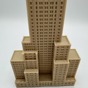 Może przedstawiać: Beżowy model architektoniczny wielopiętrowego budynku. Model przedstawia centralną wieżę otoczoną niższymi strukturami, wszystkie z licznymi prostokątnymi oknami. Fasada budynku ma szczeg&oacute;łowy, teksturowany wygląd.