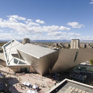May include: An exterior view of a modern art museum with a unique, angular design. The building is made of metal and glass and has a large, open space in the center. The museum is surrounded by a city skyline and a blue sky with white clouds.