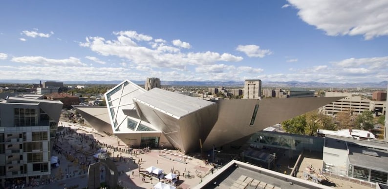 May include: An exterior view of a modern art museum with a unique, angular design. The building is made of metal and glass and has a large, open space in the center. The museum is surrounded by a city skyline and a blue sky with white clouds.