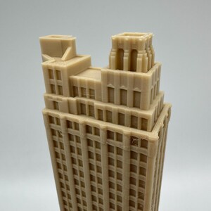 May include: A tan-colored, miniature skyscraper model with a detailed facade. The building has a tiered design with multiple windows and architectural details. The model is a representation of a classic building.