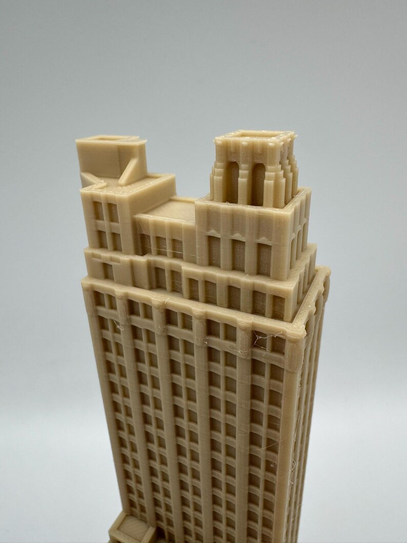 May include: A tan-colored, miniature skyscraper model with a detailed facade. The building has a tiered design with multiple windows and architectural details. The model is a representation of a classic building.