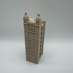 Può includere: Un modello architettonico di grattacielo stampato in 3D color beige chiaro, caratterizzato da una struttura rettangolare con una griglia di finestre, un tetto piatto e due torri a cupola prominenti sugli angoli superiori, con dettagli di facciata.