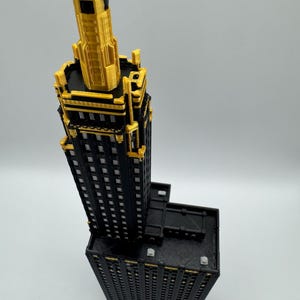 Op de afbeelding: Een gedetailleerd 3D-geprint model van een wolkenkrabber. Het gebouw heeft een zwarte gevel met gouden accenten en een gouden torenspits. Het model staat tegen een effen achtergrond.