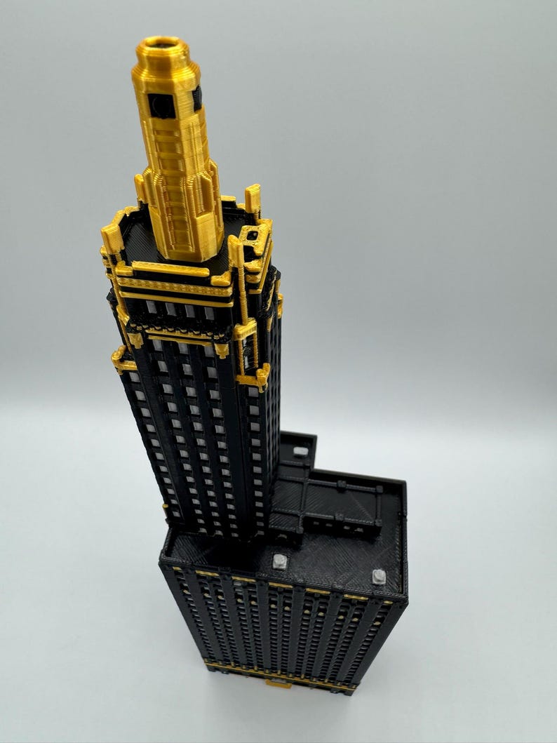 Op de afbeelding: Een gedetailleerd 3D-geprint model van een wolkenkrabber. Het gebouw heeft een zwarte gevel met gouden accenten en een gouden torenspits. Het model staat tegen een effen achtergrond.