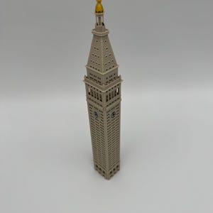 May include: A beige architectural model of the Metropolitan Life Insurance Company Tower. The structure features a square base, multiple levels of windows, a clock face on the upper section, and a pointed spire topped with a gold-colored finial.