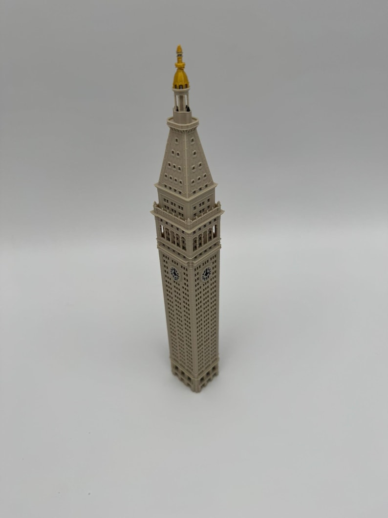 May include: A beige architectural model of the Metropolitan Life Insurance Company Tower. The structure features a square base, multiple levels of windows, a clock face on the upper section, and a pointed spire topped with a gold-colored finial.