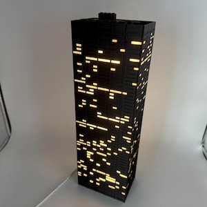 Peut inclure: Une lampe noire en forme de gratte-ciel avec des fen&ecirc;tres &eacute;clair&eacute;es. La lampe est rectangulaire et a une surface textur&eacute;e. Les fen&ecirc;tres &eacute;mettent une lumi&egrave;re jaune chaude, cr&eacute;ant un effet de paysage urbain. La lampe est branch&eacute;e avec un cordon blanc.