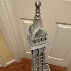 May include: A silver-colored model of the Eiffel Tower. The tower is intricately detailed, with a lattice-like structure and architectural features. The model is standing on a brown carpet in front of a white door.